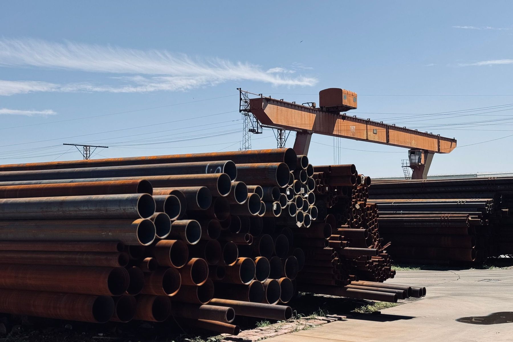 Stacks of steel pipes in an outdoor storage yard beneath an orange gantry crane